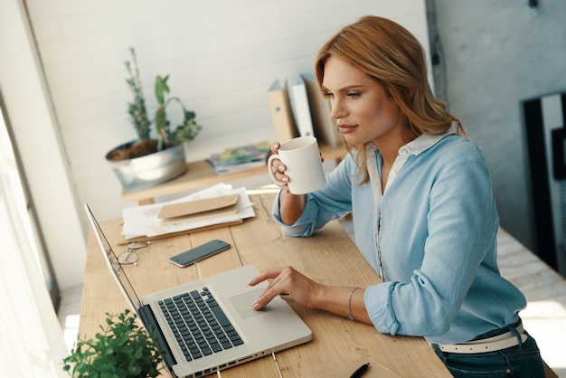 Mooie jonge vrouw in slimme vrijetijdskleding die laptop gebruikt en koffie drinkt