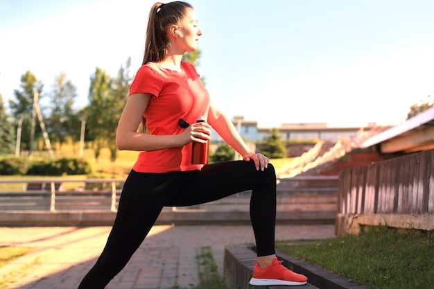 Mooie jonge vrouw in sportkleding die rekoefeningen doet in de buurt van de rivieroever van de stad, met een fles water.