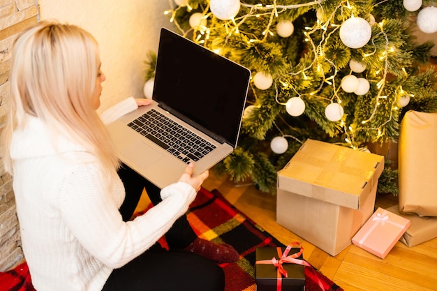 Mooie jonge vrouw in trendy wintermode zittend op de vloer met gekruiste benen met laptopgadget vooraan kerstboomversiering, terwijl ze naar de camera kijkt.