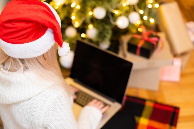 Mooie jonge vrouw in trendy wintermode zittend op de vloer met gekruiste benen met laptopgadget vooraan kerstboomversiering, terwijl ze naar de camera kijkt.