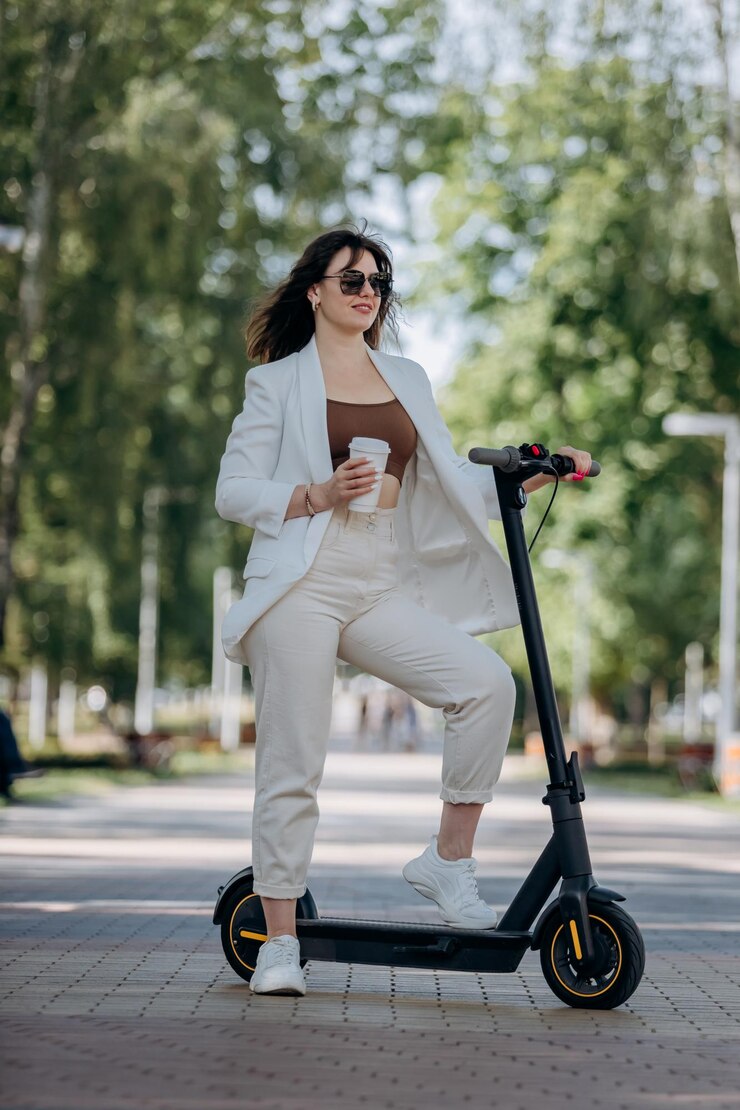 Mooie jonge vrouw in wit pak en zonnebril die met haar elektrische scooter in het stadspark staat en koffie drinkt