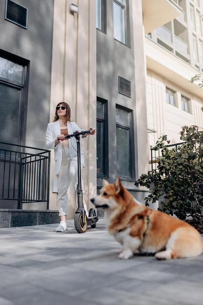 Mooie jonge vrouw in wit pak staat met haar elektrische scooter en Corgi Dog in de buurt van modern gebouw in de stad en kijkt weg