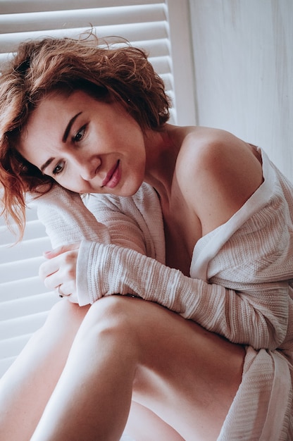 Foto mooie jonge vrouw in witte blouse draagt schouderbenen zittend op het raam in de ochtendontspanning