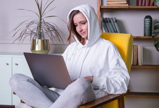 Mooie jonge vrouw in witte hoodie met laptop zittend op gele fauteuil thuis