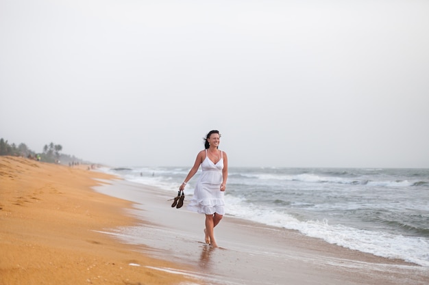 Mooie jonge vrouw in witte jurk lopen op het zandstrand, met sandalen. reizen en zomer concept