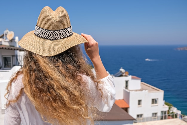 Mooie jonge vrouw in witte jurk strooien hoed op wit terras balkon van huis of hotel met uitzicht op zee.