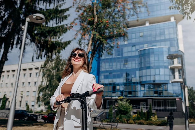 Mooie jonge vrouw in zonnebril en wit pak staande op haar elektrische scooter in de buurt van modern gebouw en wegkijkend