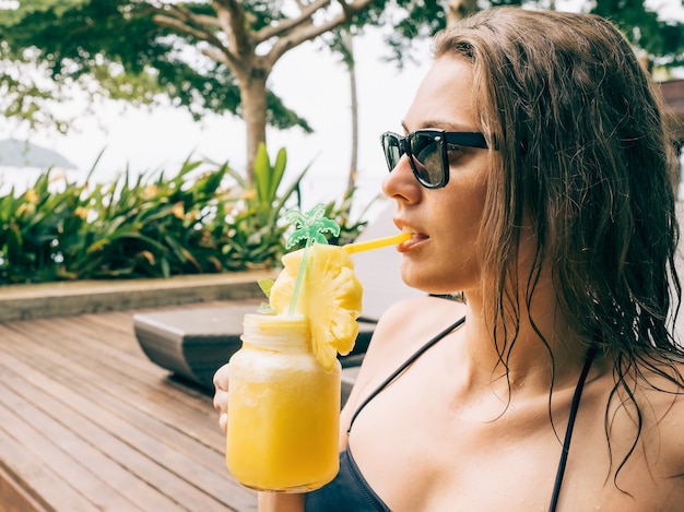 Mooie jonge vrouw in zwarte bikini cocktail drinken terwijl u ontspant in de zonnebank bij het zwembad