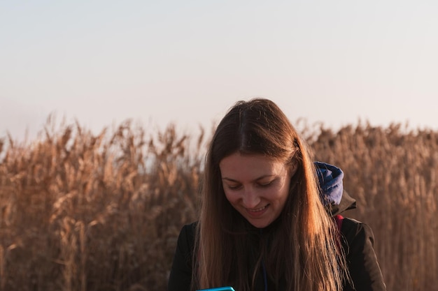 Mooie jonge vrouw in zwarte jas met rugzak smartphone op pampas gras zonsondergang hemel droog riet Millennial lang haar meisje wandelen tijd alleen doorbrengen Lifestyle echte mensen met mobiel apparaat buiten