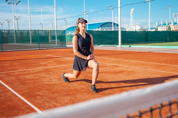 Mooie jonge vrouw in zwarte sportkleding strekt zich uit op de tennisbaan