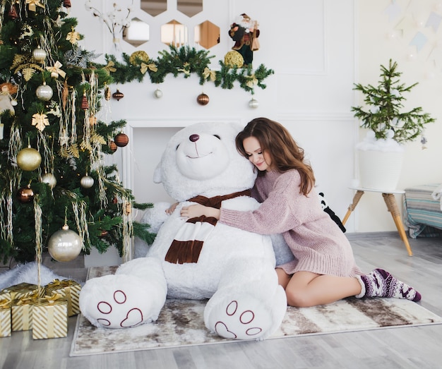 Mooie jonge vrouw knuffelen enorme teddybeer in de buurt van de kerstboom