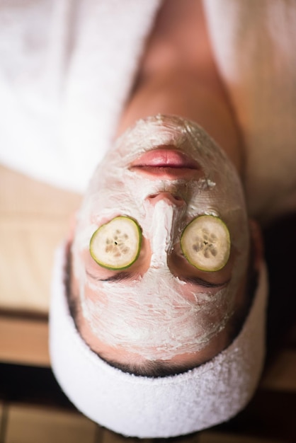 Mooie jonge vrouw krijgt een gezichtsmasker van klei in de spa, liggend met komkommers op de ogen