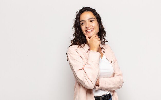 mooie jonge vrouw lachend met een gelukkige, zelfverzekerde uitdrukking met hand op de kin, zich afvragend en opzij kijkend