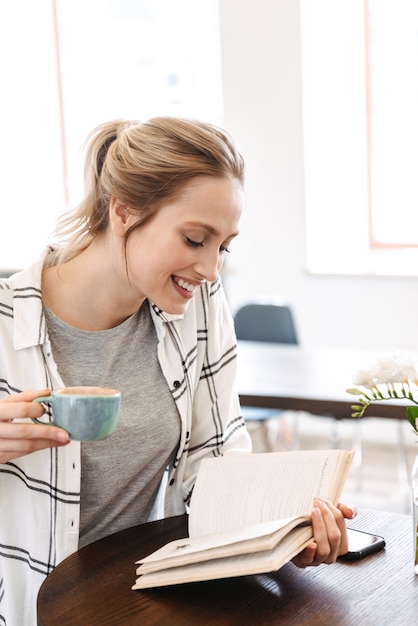 Mooie jonge vrouw leesboek zittend binnenshuis koffie drinken, beker te houden