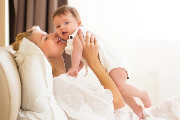 Mooie jonge vrouw met babymeisje in haar armen op de binnenlandse kamer in de ochtend. Gelukkige moeder knuffelt en kust haar kleine pasgeboren dochter thuis op wit bed