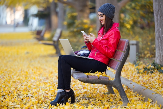 Mooie jonge vrouw met behulp van haar mobiele telefoon tijdens het werken met laptop in de herfst.