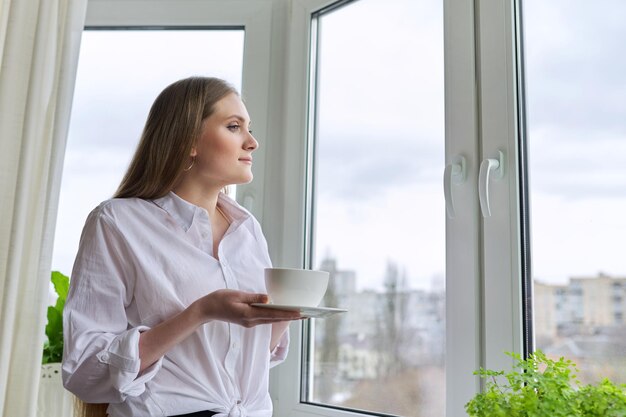 Mooie jonge vrouw met blond lang haar met kopje koffie in de buurt van raam