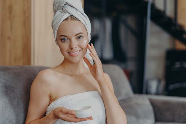 Mooie jonge vrouw met een gezonde gladde huid past gezichtscrème toe, draagt een gewikkelde handdoek op het hoofd na het nemen van een douche, vormt op comfortabele bank.
