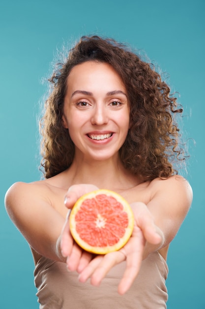 Mooie jonge vrouw met een gezonde huid en een brede glimlach die je de helft van verse, sappige grapefruit laat zien of geeft terwijl je het in handen houdt