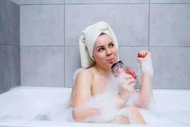 Mooie jonge vrouw met een handdoek op haar hoofd die een cocktail drinkt, neemt thuis een bad. ontspan na een zware dag. de spa is een ontspannende procedure.