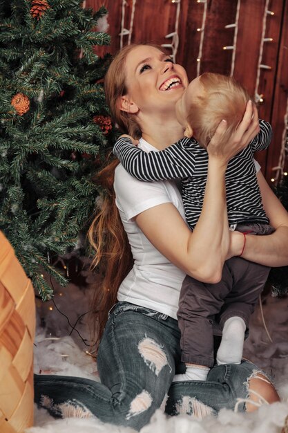 Mooie jonge vrouw met een jaar oud kind dat bij de kerstboom in de woonkamer speelt. Moeder met schattige zoon in kerst ingerichte kamer. Ze glimlachen en zijn blij. Concept van familie Nieuwjaar. Ruimte kopiëren