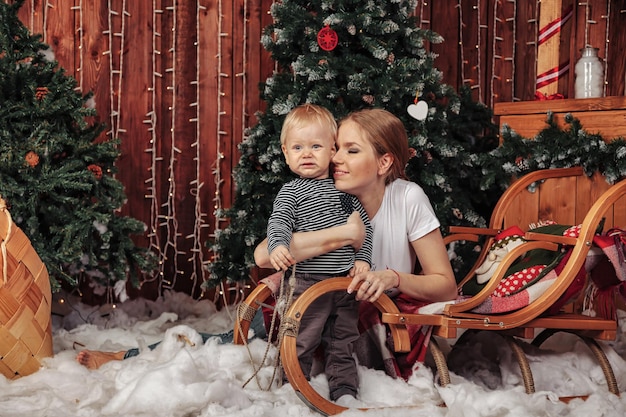 Mooie jonge vrouw met een jaar oud kind dat bij de kerstboom in de woonkamer speelt. Moeder met schattige zoon in kerst ingerichte kamer. Ze glimlachen en zijn blij. Concept van familie Nieuwjaar. Ruimte kopiëren