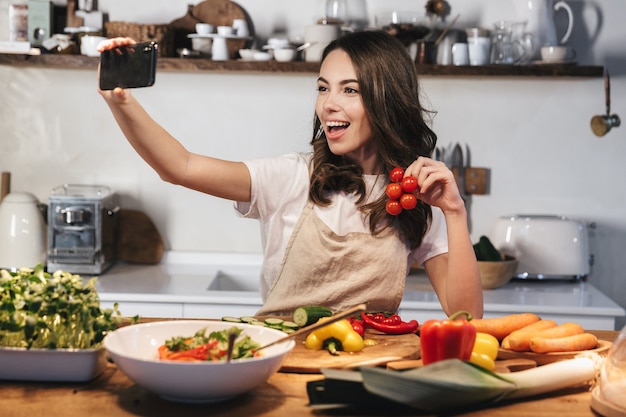 Mooie jonge vrouw met een schort die een gezonde salade kookt in de keuken thuis, een selfie maakt met mobiele telefoon