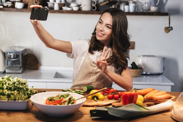 Mooie jonge vrouw met een schort die een gezonde salade kookt in de keuken thuis, een selfie maakt met mobiele telefoon