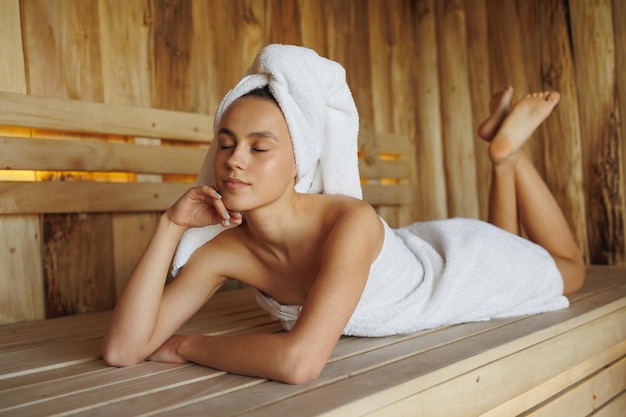 Foto mooie jonge vrouw met gesloten ogen die in een houten sauna ligt.