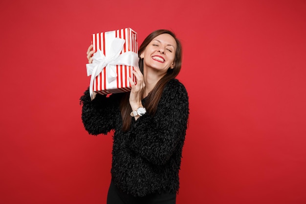 Mooie jonge vrouw met gesloten ogen houdt rood gestreepte huidige doos met cadeaulint geïsoleerd op rode achtergrond. Valentijnsdag, Internationale Vrouwendag, verjaardag, vakantieconcept. Bespotten kopie ruimte.