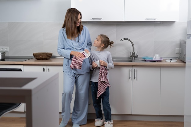 Mooie jonge vrouw met haar dochter in de keuken veegt de afwas af, kijkt elkaar aan en glimlacht terwijl ze de keuken schoonmaakt