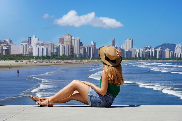 Mooie jonge vrouw met hoed zittend op de grond met skyline van de metropool en golvende oceaan op onscherpe achtergrond