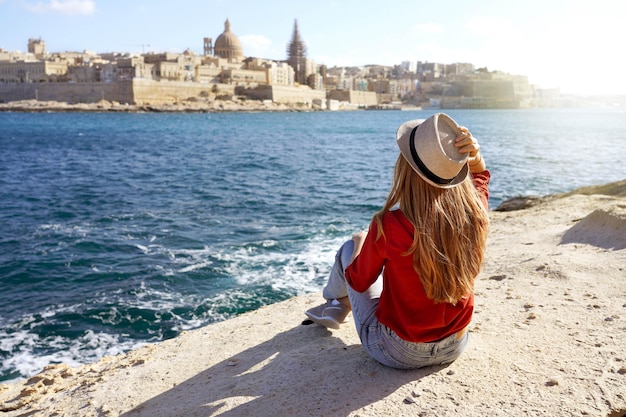 Mooie jonge vrouw met hoed zittend op steen aan zee en kijkend naar een prachtig panoramisch uitzicht over de stad Valletta in Malta