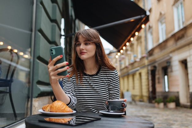 Mooie jonge vrouw met koffie zittend aan café tafel en kijken naar mobiel scherm