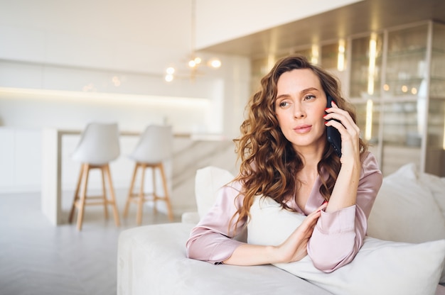 Mooie jonge vrouw met krullend haar in roze pyjama's op witte bank in de ochtend. Dame sprekende telefoon in Scandinavische stijl woonkamer interieur.