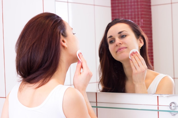 Mooie jonge vrouw met lang donker haar in wit overhemd dat make-up verwijdert met wattenschijfje voor de spiegel in haar badkamer