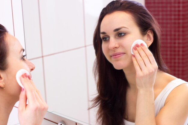 Mooie jonge vrouw met lang donker haar in wit overhemd die make-up verwijdert met wattenschijfje voor de spiegel in haar badkamer