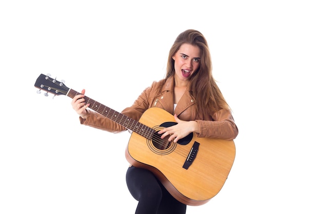 Mooie jonge vrouw met lang haar in bruine jas en zwarte broek die een gitaar vasthoudt en lacht op een witte achtergrond in de studio