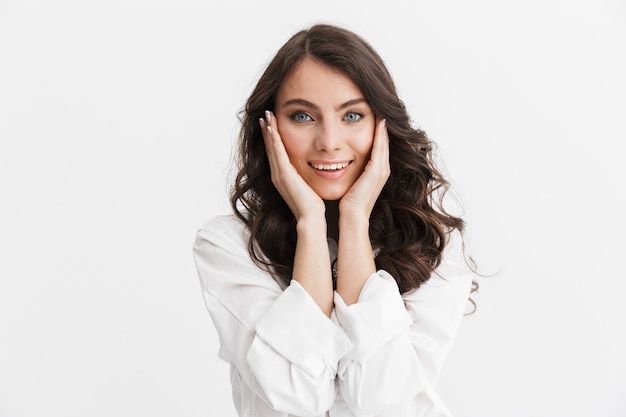 Mooie jonge vrouw met lang krullend donkerbruin haar, gekleed in een wit overhemd, geïsoleerd over een witte muur