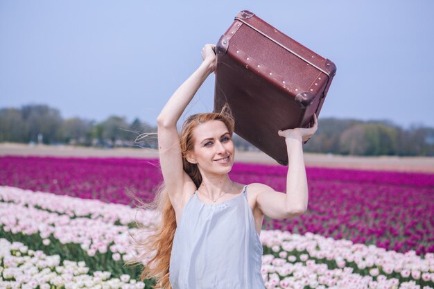 Mooie jonge vrouw met lang rood haar dat in witte kleding draagt die zich met bagage op kleurrijk tulpengebied bevindt.