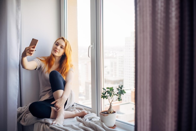 Mooie jonge vrouw met lang rood haar neemt een selfie op haar smartphone terwijl ze op de vensterbank zit. Mooi meisje met een telefoon in een thuisomgeving. Technologie en sociale netwerken concept.