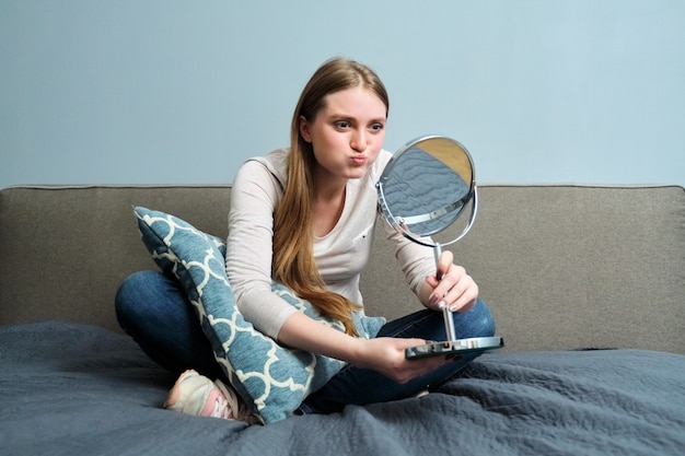 Mooie jonge vrouw met make-up spiegel om thuis in bed te zitten, meisje maakt emoties, kijkt naar zichzelf in spiegel