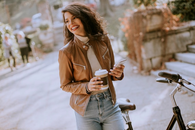 Mooie jonge vrouw met mobiele telefoon drinkt koffie om met de fiets te gaan op herfstdag