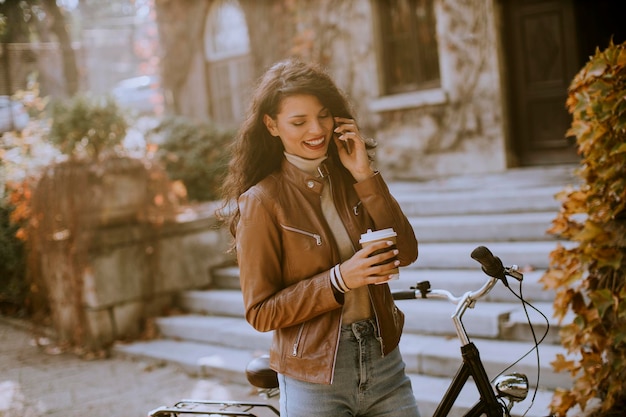 Mooie jonge vrouw met mobiele telefoon drinkt koffie om met de fiets te gaan op herfstdag