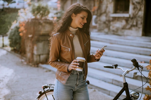 Mooie jonge vrouw met mobiele telefoon drinkt koffie om met de fiets te gaan op herfstdag