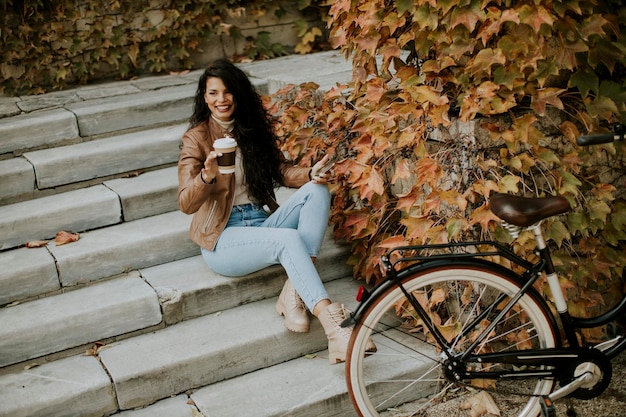 Mooie jonge vrouw met mobiele telefoon drinkt koffie om op herfstdag met de fiets naar de trap te gaan