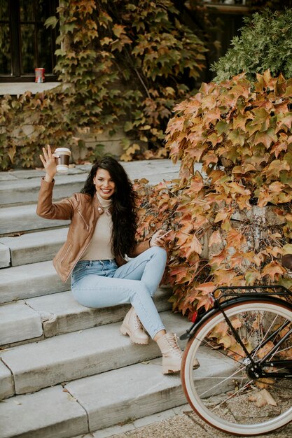 Mooie jonge vrouw met mobiele telefoon drinkt koffie om op herfstdag met de fiets naar de trap te gaan