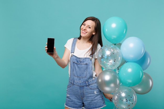 Mooie jonge vrouw met mobiele telefoon met leeg leeg scherm, vieren met kleurrijke lucht ballonnen geïsoleerd op blauwe turquoise muur achtergrond. vakantie verjaardagsfeestje, mensen emoties concept.