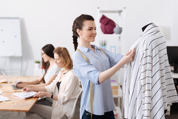 Mooie jonge vrouw met revers van een vest die aan een mannequin hangt terwijl haar collega's outfitschetsen ontwikkelen