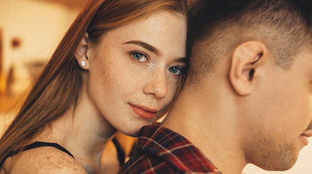 Mooie jonge vrouw met rood haar en sproeten glimlachen terwijl het omhelzen van achter haar vriendje thuis in de keuken.